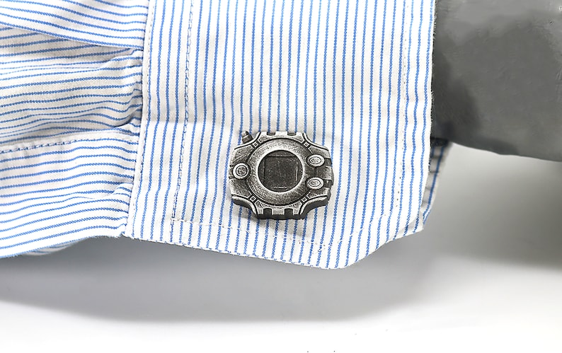 May include: A pair of silver cufflinks featuring a detailed design of a circular device with a square window in the center.