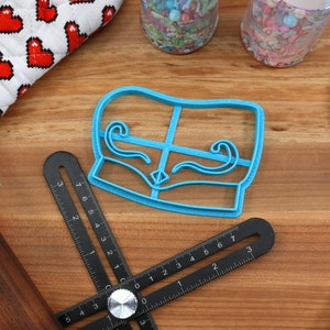 May include: A light blue cookie cutter in the shape of a couch with decorative swirls, a black adjustable ruler, a wooden rolling pin, and a blue spatula are on a wooden surface. A heart-patterned oven mitt and jars of sprinkles are also visible.