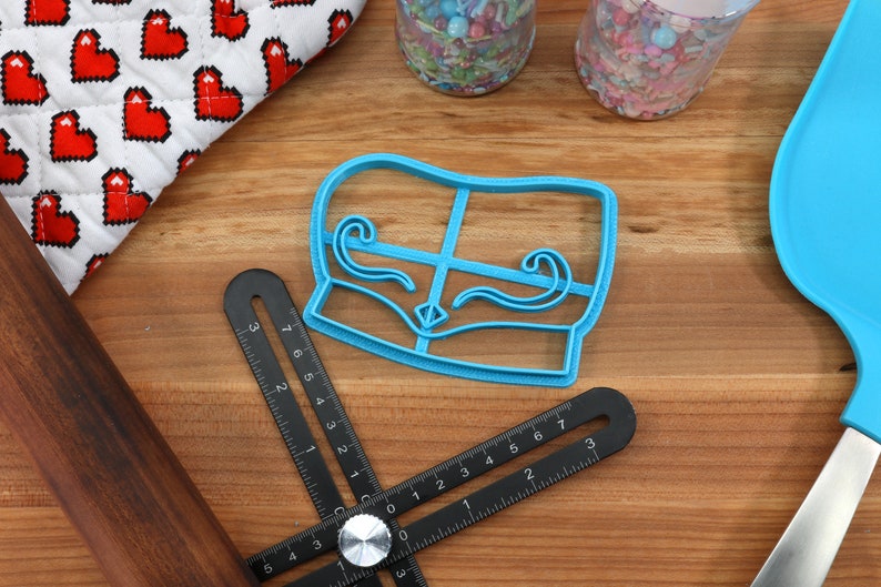 May include: A light blue cookie cutter in the shape of a couch with decorative swirls, a black adjustable ruler, a wooden rolling pin, and a blue spatula are on a wooden surface. A heart-patterned oven mitt and jars of sprinkles are also visible.
