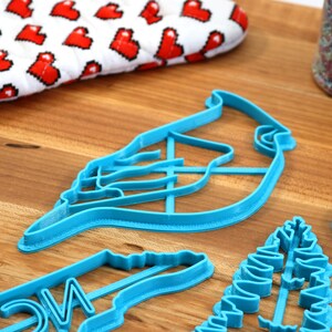 May include: A set of four blue plastic cookie cutters shaped like a cardinal, a pine branch, a Christmas tree, and a wreath. The cutters are on a wooden surface.