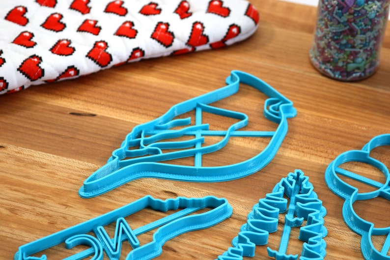 May include: A set of four blue plastic cookie cutters shaped like a cardinal, a pine branch, a Christmas tree, and a wreath. The cutters are on a wooden surface.