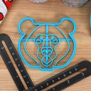 May include: A blue plastic bear-shaped cookie cutter. The cutter is about 2 inches in diameter and has a detailed design of a bear's face.