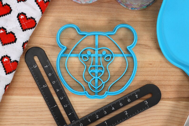 May include: A blue plastic bear-shaped cookie cutter. The cutter is about 2 inches in diameter and has a detailed design of a bear's face.