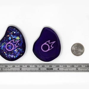 May include: Two purple and blue resin cabochons with a pink and white fire symbol. The cabochons are approximately 2 inches in diameter. A US quarter is shown for scale.