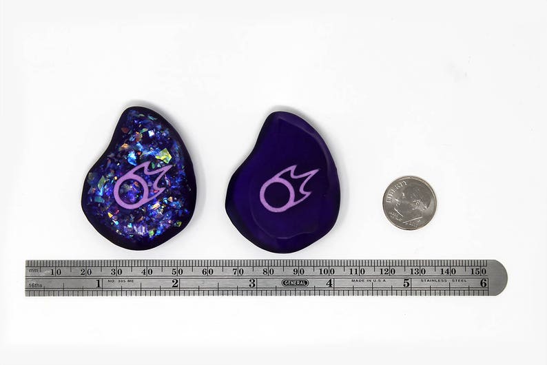 May include: Two purple and blue resin cabochons with a pink and white fire symbol. The cabochons are approximately 2 inches in diameter. A US quarter is shown for scale.