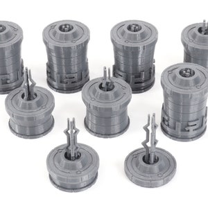 May include: Eight gray 3D printed miniature towers with intricate designs. The towers are cylindrical with a circular base and a pointed top. The towers are arranged in a random pattern on a white surface.