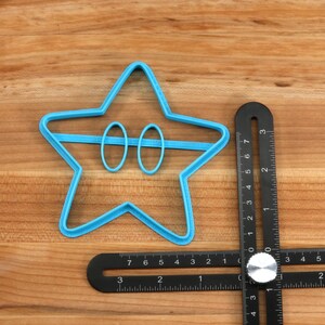 May include: A blue plastic star-shaped cookie cutter with two circles cut out in the center. The cutter is on a wooden surface next to a black metal ruler.