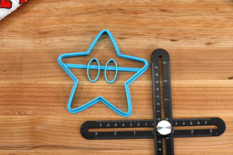 May include: A blue plastic star-shaped cookie cutter with two circles cut out in the center. The cutter is on a wooden surface next to a black metal ruler.