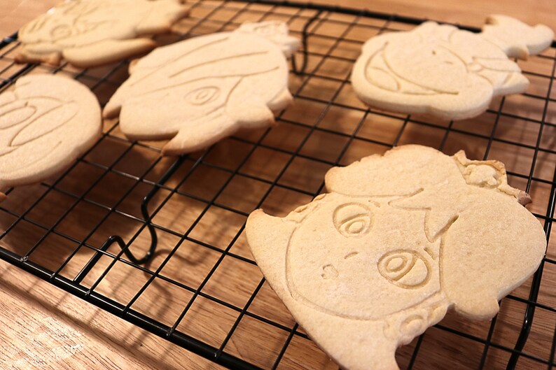 May include: Four sugar cookies decorated with icing and designs of cartoon characters on a black wire cooling rack.