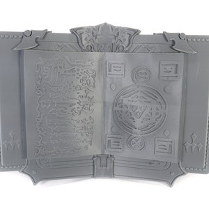May include: A gray, 3D printed book with an intricate design on the cover and pages. The book is open to reveal a detailed, circular pattern on the right page and a more abstract design on the left page.