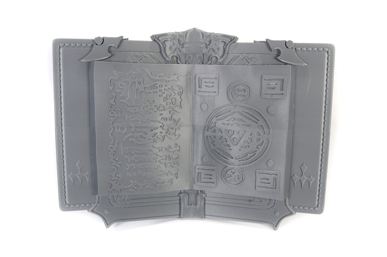 May include: A gray, 3D printed book with an intricate design on the cover and pages. The book is open to reveal a detailed, circular pattern on the right page and a more abstract design on the left page.