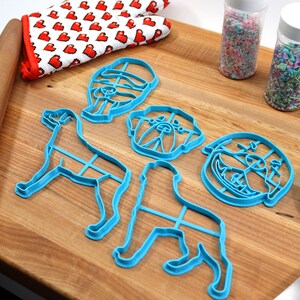 May include: A set of five blue plastic dog-shaped cookie cutters on a wooden cutting board. The cutters include a full dog, a dog head, and three different dog head expressions. There are also two jars of sprinkles and a blue spatula.