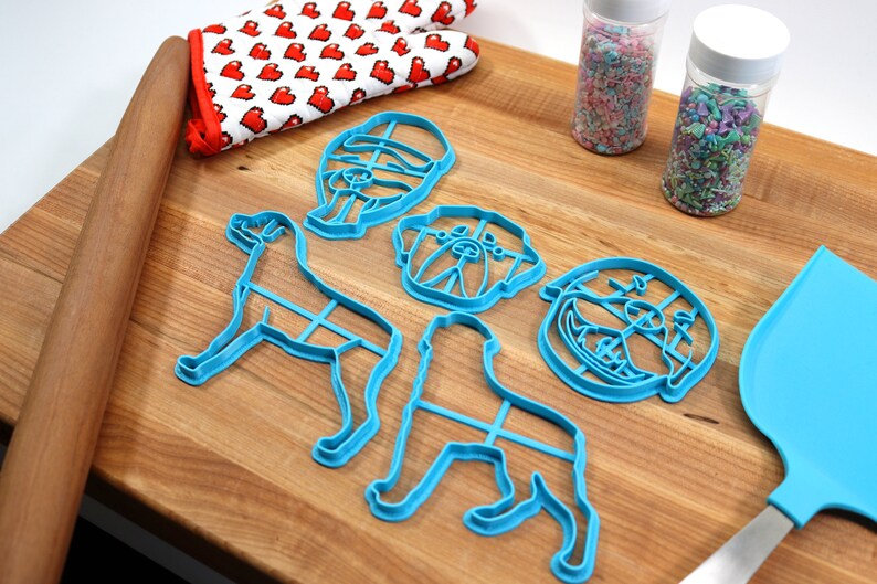 May include: A set of five blue plastic dog-shaped cookie cutters on a wooden cutting board. The cutters include a full dog, a dog head, and three different dog head expressions. There are also two jars of sprinkles and a blue spatula.