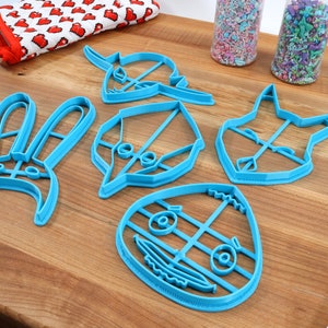 May include: Five blue plastic cookie cutters shaped like a bunny, a leaf, a cat, a character with a large head and eyes, and a character with a large head and a smile. The cutters are on a wooden surface.