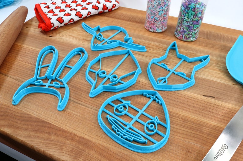 May include: Five blue plastic cookie cutters shaped like a bunny, a leaf, a cat, a character with a large head and eyes, and a character with a large head and a smile. The cutters are on a wooden surface.