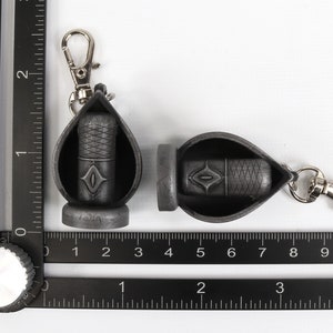 May include: Two black keychains with a silver clip. Each keychain has a small black pouch with a diamond-shaped design. The pouches are open and appear to hold a small black object.