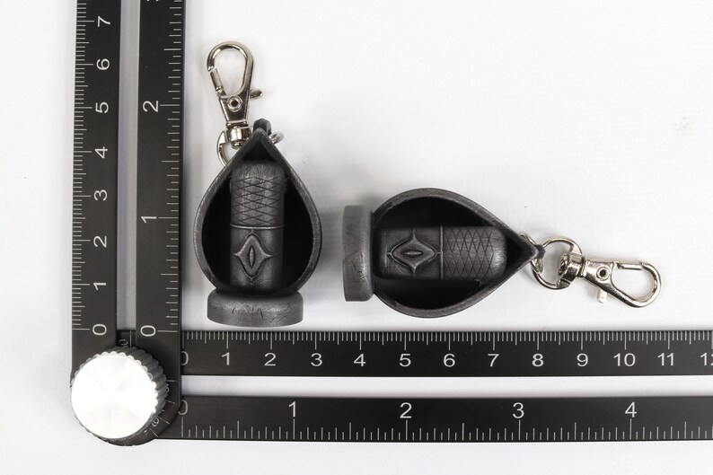 May include: Two black keychains with a silver clip. Each keychain has a small black pouch with a diamond-shaped design. The pouches are open and appear to hold a small black object.