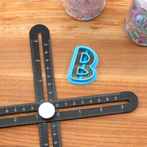 Op de afbeelding: Een blauwe plastic letter B koekjesvormer op een houten oppervlak met een zwarte liniaal. De liniaal heeft markeringen in inches.