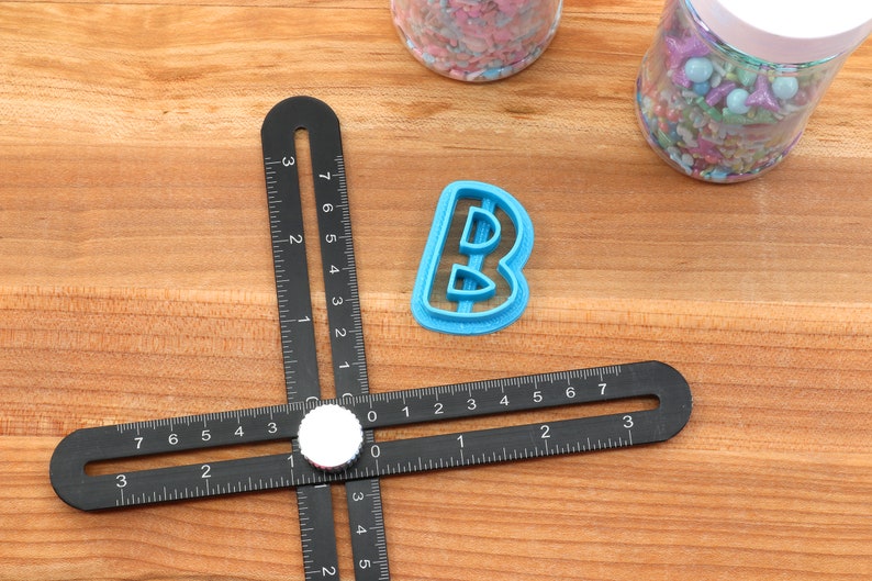 Op de afbeelding: Een blauwe plastic letter B koekjesvormer op een houten oppervlak met een zwarte liniaal. De liniaal heeft markeringen in inches.