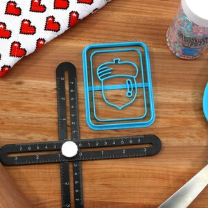 May include: A blue plastic acorn-shaped cookie cutter on a wooden surface with a black ruler, a wooden rolling pin, a blue spatula, and a jar of sprinkles.