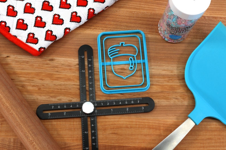 May include: A blue plastic acorn-shaped cookie cutter on a wooden surface with a black ruler, a wooden rolling pin, a blue spatula, and a jar of sprinkles.