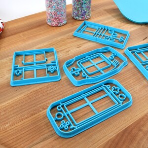 May include: Five blue plastic cookie cutters shaped like different handheld video game consoles. The cutters are on a wooden surface.