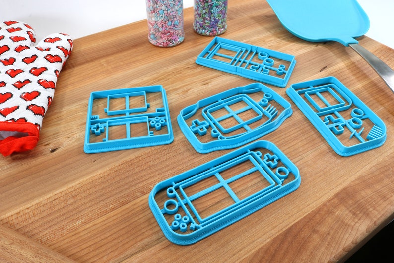May include: Five blue plastic cookie cutters shaped like different handheld video game consoles. The cutters are on a wooden surface.