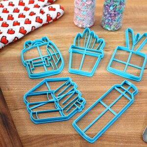 K&ouml;nnte beinhalten: Ein Set mit sechs blauen Plastik-Ausstechern in Form eines Rucksacks, einer Topfpflanze, einer Geschenkbox, eines Stapels B&uuml;cher und einer Wasserflasche. Die Ausstecher befinden sich auf einer Holzoberfl&auml;che.