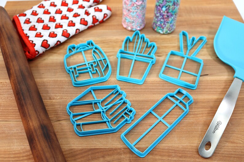 K&ouml;nnte beinhalten: Ein Set mit sechs blauen Plastik-Ausstechern in Form eines Rucksacks, einer Topfpflanze, einer Geschenkbox, eines Stapels B&uuml;cher und einer Wasserflasche. Die Ausstecher befinden sich auf einer Holzoberfl&auml;che.