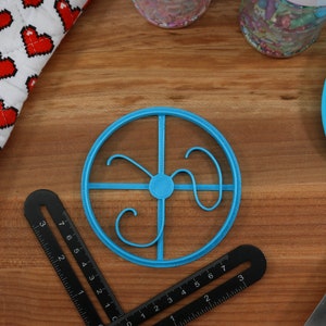 Op de afbeelding: Een blauwe plastic koekjesvormer in de vorm van een cirkel met een decoratief ontwerp erin. De vormer ligt op een houten oppervlak.