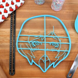 May include: A light blue plastic cookie cutter shaped like a cartoon face with a grid pattern. The cookie cutter is on a wooden surface.