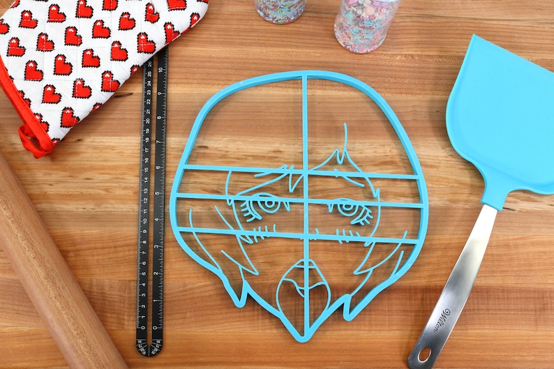May include: A light blue plastic cookie cutter shaped like a cartoon face with a grid pattern. The cookie cutter is on a wooden surface.
