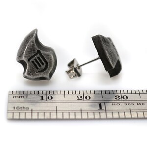 May include: A pair of dark gray metal stud earrings. Each earring has a unique, abstract shape with a lyre symbol etched into the surface. The earrings are shown with a ruler, with measurements in inches.