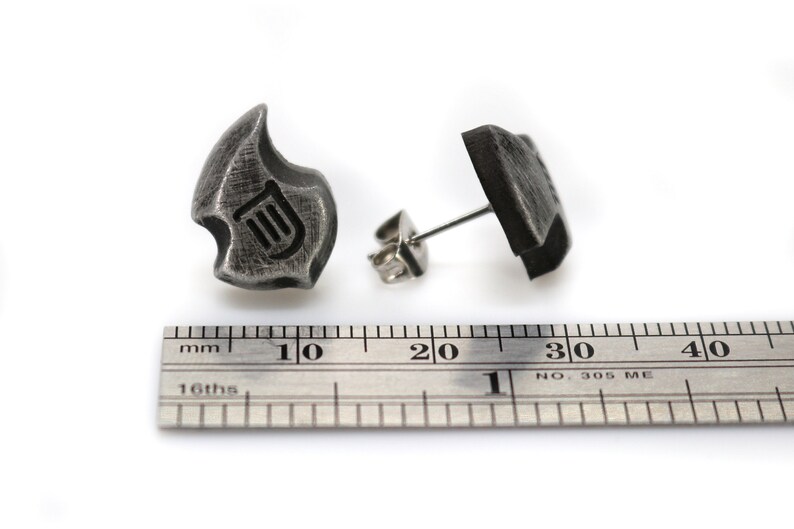May include: A pair of dark gray metal stud earrings. Each earring has a unique, abstract shape with a lyre symbol etched into the surface. The earrings are shown with a ruler, with measurements in inches.