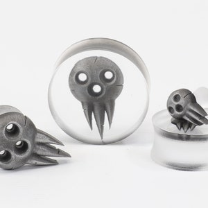 May include: A collection of gray skull-shaped ear gauges and plugs. The designs feature three circular eye holes and a jagged mouth. One plug is encased in clear acrylic, while others are standalone.