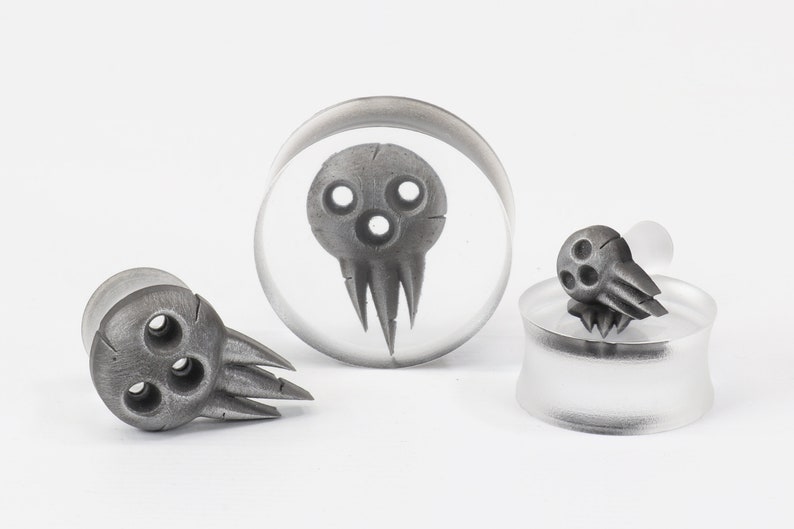May include: A collection of gray skull-shaped ear gauges and plugs. The designs feature three circular eye holes and a jagged mouth. One plug is encased in clear acrylic, while others are standalone.