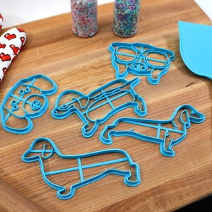 May include: Five blue plastic cookie cutters shaped like dachshunds, one with a pair of glasses, on a wooden cutting board. There is a rolling pin, a red oven mitt, and a blue spatula with a silver handle.