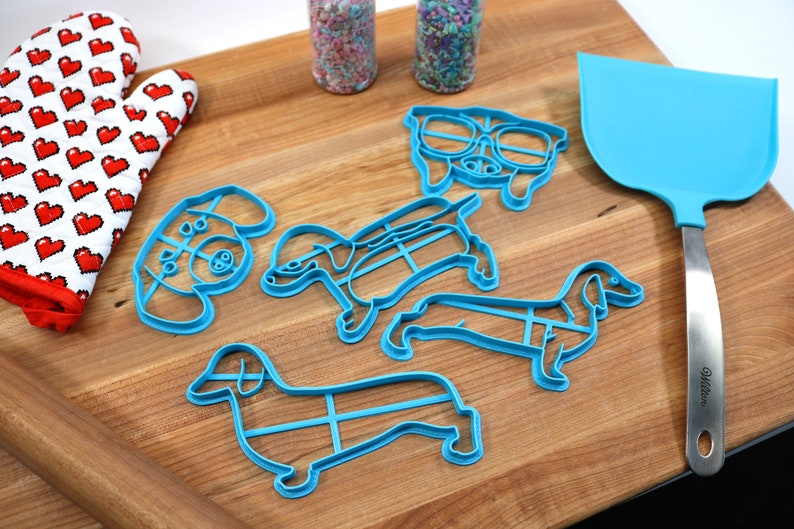 May include: Five blue plastic cookie cutters shaped like dachshunds, one with a pair of glasses, on a wooden cutting board. There is a rolling pin, a red oven mitt, and a blue spatula with a silver handle.