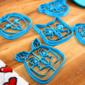 May include: Five blue plastic cookie cutters shaped like animal faces. The cutters are on a wooden surface.