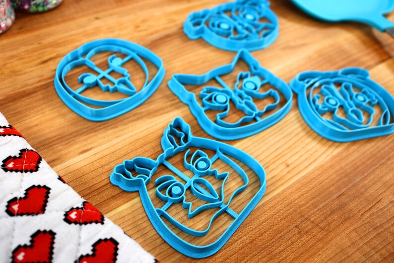 May include: Five blue plastic cookie cutters shaped like animal faces. The cutters are on a wooden surface.