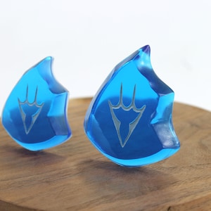 May include: Two blue crystal-like pendants with a white design in the shape of a stylized hand. The pendants are on a wooden surface.