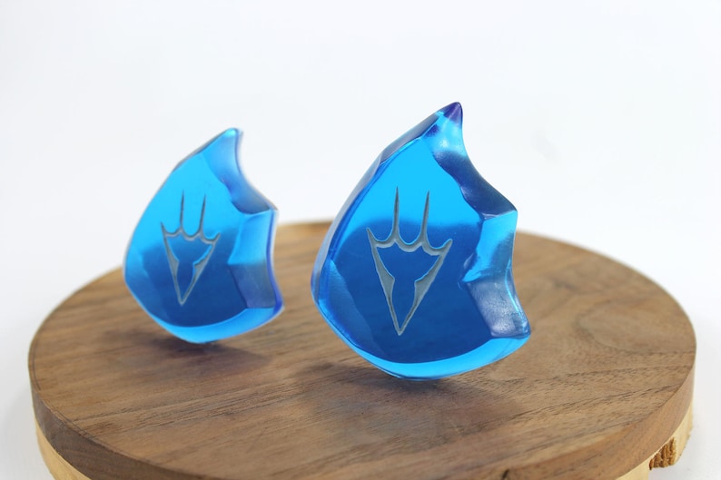 May include: Two blue crystal-like pendants with a white design in the shape of a stylized hand. The pendants are on a wooden surface.