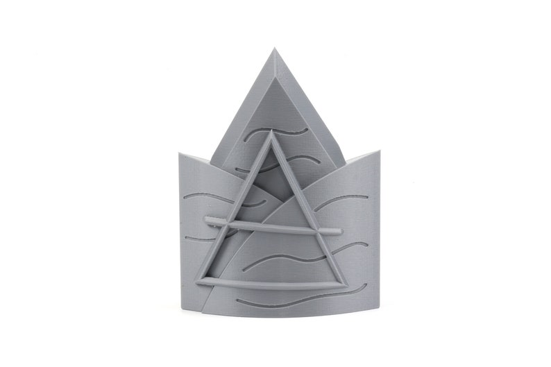May include: A gray 3D printed crown with a triangle design and wavy lines. The crown is made of plastic and has a smooth finish.