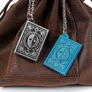Puede incluir: Dos colgantes plateados y azules con un dise&ntilde;o de cara colgando de cadenas en una bolsa de cuero marr&oacute;n. Los colgantes tienen forma de cartas de juego.