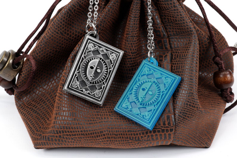 Puede incluir: Dos colgantes plateados y azules con un dise&ntilde;o de cara colgando de cadenas en una bolsa de cuero marr&oacute;n. Los colgantes tienen forma de cartas de juego.