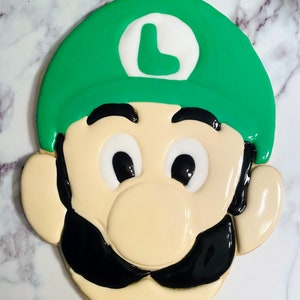 May include: A decorated sugar cookie in the shape of Luigi from the Super Mario Bros. video game series. The cookie features a green hat with a white circle and a green "L", black mustache, and black eyes. The cookie is beige with white and black icing.