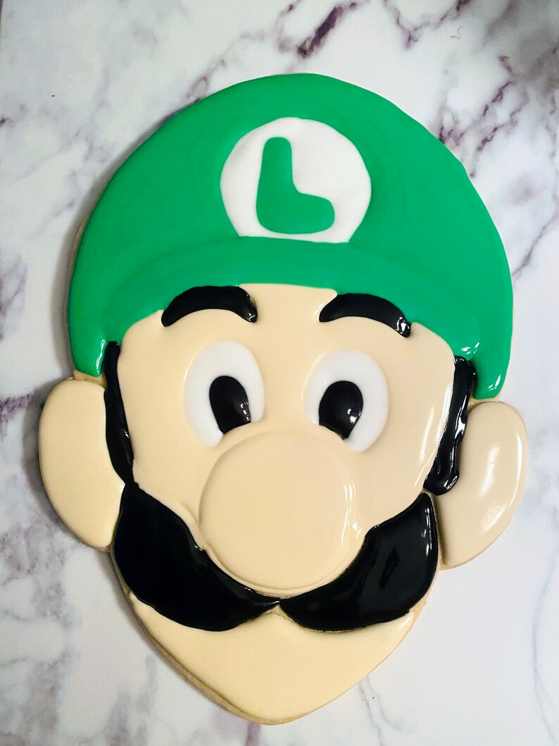 May include: A decorated sugar cookie in the shape of Luigi from the Super Mario Bros. video game series. The cookie features a green hat with a white circle and a green "L", black mustache, and black eyes. The cookie is beige with white and black icing.