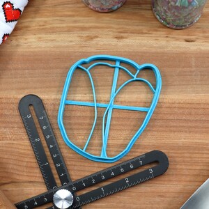 May include: A blue plastic cookie cutter shaped like a heart with a lattice design. The cutter is on a wooden surface with a ruler and a rolling pin.