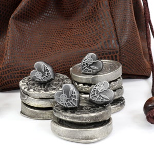 May include: A set of silver heart-shaped tokens with a web design, stacked in piles of two and three, next to a brown leather pouch with a drawstring.
