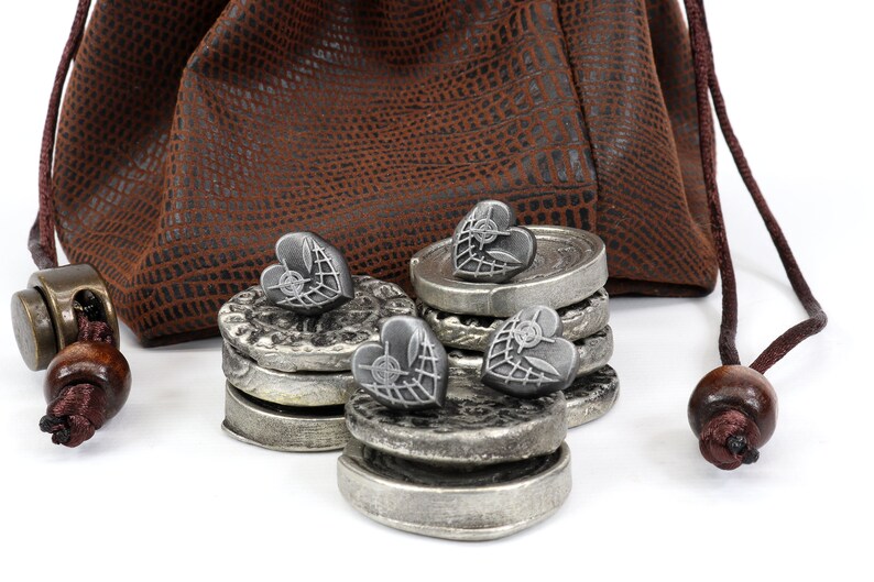 May include: A set of silver heart-shaped tokens with a web design, stacked in piles of two and three, next to a brown leather pouch with a drawstring.